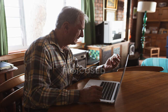 Vista lateral de um homem caucasiano sênior relaxando em casa em sua sala de estar, sentado à mesa usando um computador portátil e segurando uma garrafa de comprimidos — Fotografia de Stock