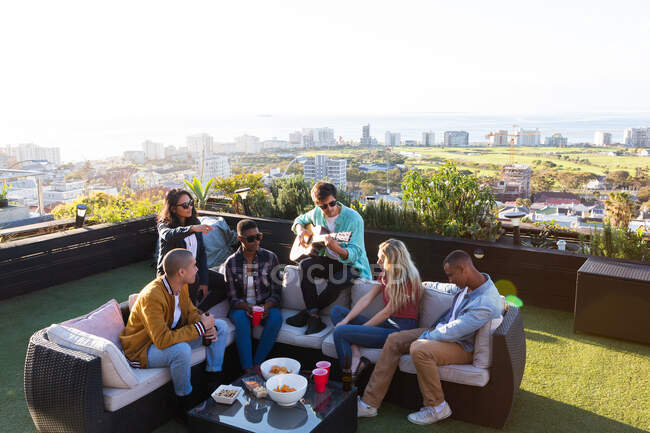 Vista de ángulo alto de un grupo multiétnico de amigos pasando el rato en una terraza en un día soleado, uno de ellos tocando la guitarra - foto de stock