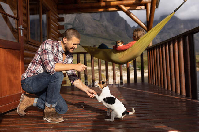 Side view of a Caucasian couple having a good trip to the mountains, a man is kneeling on a balcony in a cabin, playing with puppies, while a woman is lying on a hammock with a puppy on her belly — Stock Photo