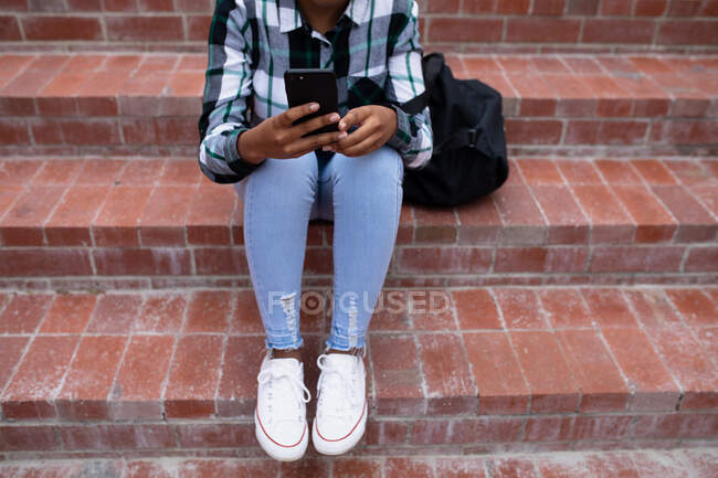 Низкий вид спереди на женщину старшеклассницу, с помощью смартфона и сидя на ступеньках на территории школы — стоковое фото