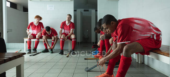 Seitenansicht einer multiethnischen Gruppe männlicher Hockeyspieler, die sich vor einem Spiel vorbereiten, in einer Umkleidekabine sitzen, sich vorbereiten, Schuhe binden und fokussieren — Stockfoto