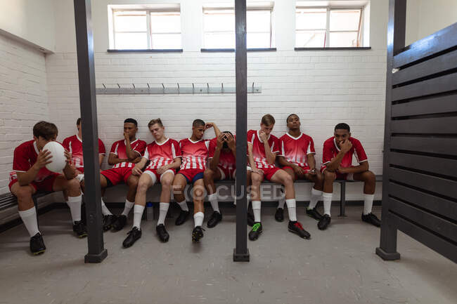 Vista frontal de un grupo de jugadores de rugby masculinos multiétnicos adolescentes con tira de equipo roja y blanca, sentados y descansando en el vestuario después de un partido - foto de stock
