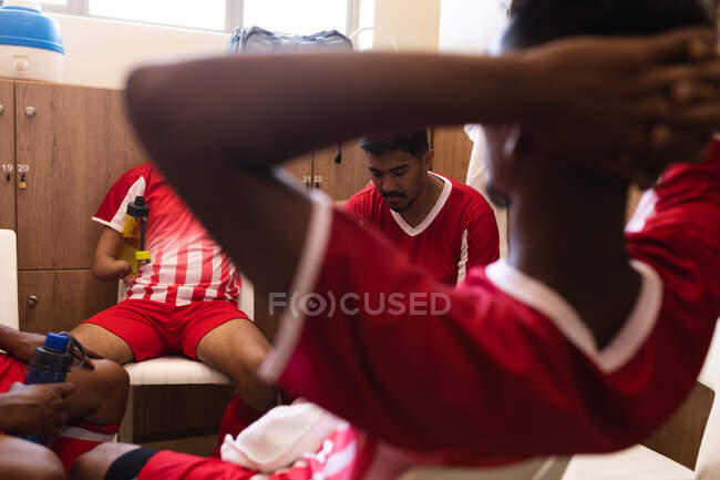 Multi ethnic group of male football players wearing a team strip sitting in changing room during a break in game, resting holding water bottles. — Stock Photo