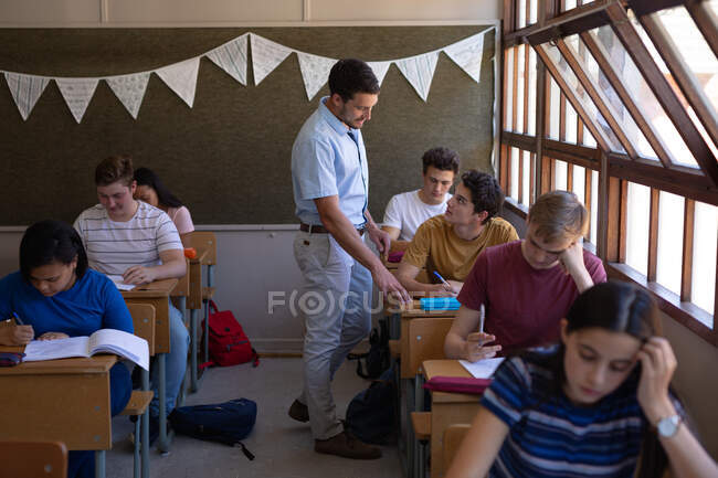 Vista frontal de un grupo multiétnico de alumnos adolescentes sentados en escritorios en clase estudiando en la escuela con un profesor caucásico de pie y hablando con un chico caucásico en su escritorio - foto de stock