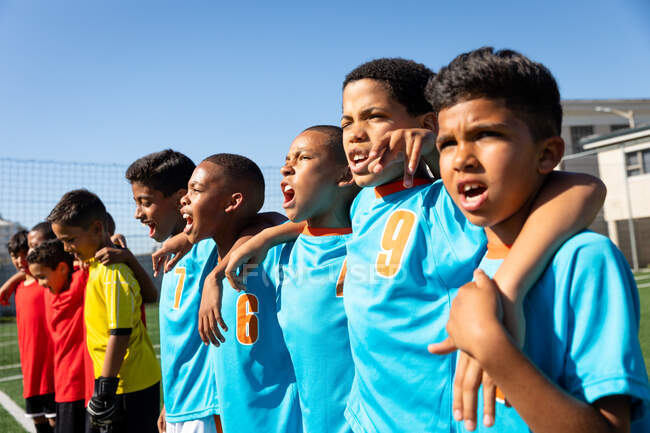Vista laterale da vicino di un gruppo multietnico di giocatori di calcio maschili che indossano strisce di squadra, in piedi su un campo di gioco, abbracciandosi con le braccia l'un l'altro e cantando — Foto stock
