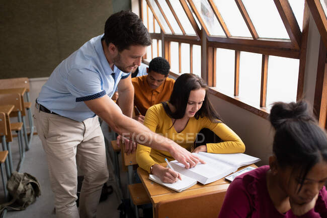 Seitenansicht eines kaukasischen männlichen Gymnasiallehrers, der mit einem jugendlichen kaukasischen Mädchen in einem Klassenzimmer auf einem Schreibtisch steht und spricht, während Teenager-Klassenkameraden an Schreibtischen im Hintergrund arbeiten. — Stockfoto