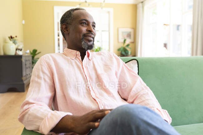 Gros plan d'un homme afro-américain à la retraite heureux et beau, assis sur le canapé dans son salon, regardant ailleurs et souriant, s'isolant lui-même pendant la pandémie de coronavirus covid19 — Photo de stock