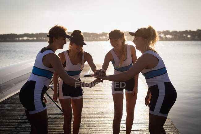 Vista frontal de un equipo de remo de cuatro mujeres caucásicas entrenando en el río, apilándose las manos y uniéndose, de pie en un embarcadero al amanecer antes de remar - foto de stock