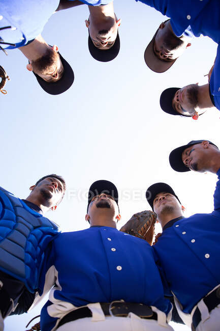 Vista de bajo ángulo de un equipo multiétnico de jugadores de béisbol masculinos, preparándose antes de un juego, motivándose mutuamente en un grupo en un día soleado - foto de stock