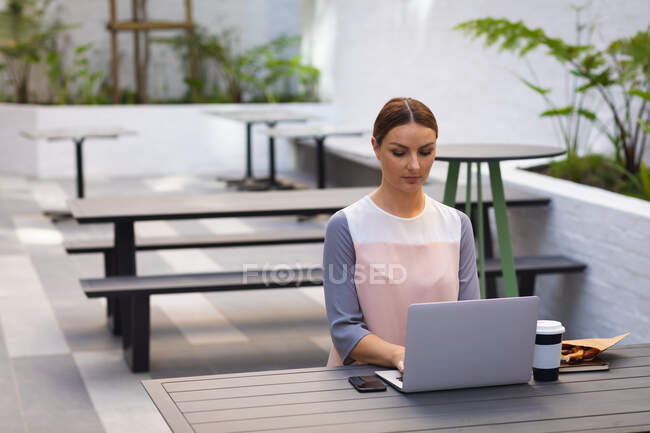 Una donna d'affari caucasica in viaggio in una giornata di sole, seduta a un tavolo e al lavoro sul suo computer portatile, con un caffè da asporto su un tavolo — Foto stock