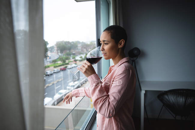 Vista lateral de uma mulher de raça mista com cabelo curto relaxante em casa, em pé na sala de estar olhando pela janela, bebendo um copo de vinho tinto — Fotografia de Stock