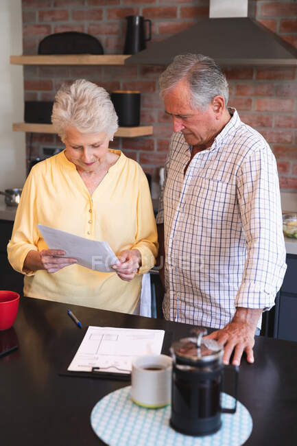 A retired senior Caucasian couple standing at a table in their dining room looking at paperwork and discussing their finances, with a pot of coffee and cup on the table, at home together isolating during coronavirus covid19 pandemic — Stock Photo