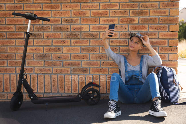 Mulher alternativa de raça mista vestindo um boné e dungarees para fora e sobre na cidade em um dia ensolarado, sentado contra a parede com sua scooter tirar uma selfie com smartphone. Nômade digital urbano em movimento . — Fotografia de Stock