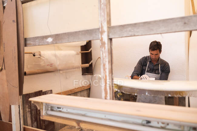Кавказский производитель досок для серфинга работает в своей студии, готовит деревянную доску для серфинга, покрытую белым куском ткани, чтобы отполировать ее и покрасить.. — стоковое фото