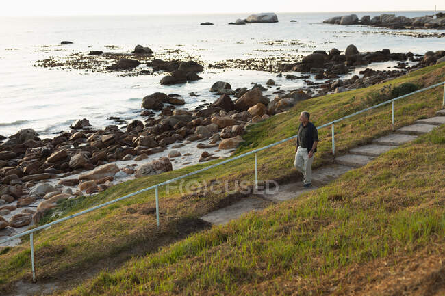 Vista lateral de un hombre mayor del Cáucaso caminando por las escaleras en el campo junto al mar, admirando una vista costera - foto de stock