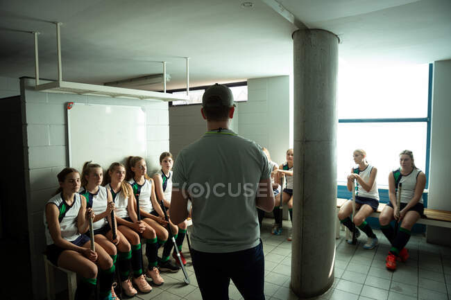 Visão traseira de um treinador de hóquei em campo caucasiano interagindo com um grupo de jogadoras de hóquei em campo caucasianas, sentadas em um vestiário — Fotografia de Stock
