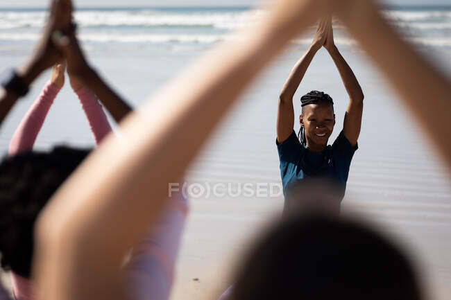 Vista frontal de una mujer atractiva afroamericana, vistiendo ropa deportiva, con los brazos en posición de yoga, de pie en la playa soleada vista con sus amigas con los brazos también levantados. - foto de stock