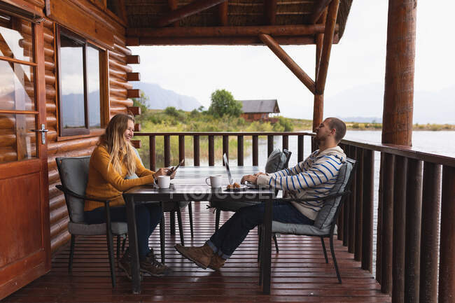 Seitenansicht eines kaukasischen Paares, das sich bei einem Ausflug in die Berge amüsiert, an einem Tisch auf einem Balkon in einer Hütte sitzt, eine Frau ihr Smartphone benutzt und ein Mann seinen Laptop — Stockfoto