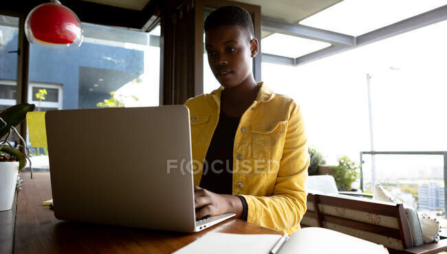 Vista frontal de una mujer afroamericana sentada en su sala de estar frente a una ventana en un día soleado, usando una computadora portátil - foto de stock
