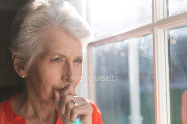 Primo piano di una donna anziana caucasica in pensione a casa seduta nel suo soggiorno in una giornata di sole, guardando fuori dalla finestra e pensando, auto isolante durante il coronavirus covid19 pandemia — Foto stock