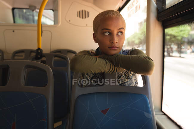 Mujer alternativa de raza mixta con pelo corto y rubio en la ciudad, sentada en un autobús, apoyada en el asiento delantero, mirando por la ventana. Nómada urbano independiente en movimiento. - foto de stock