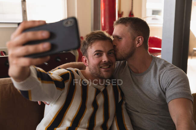 Vista frontal de cerca de la pareja masculina caucásica relajándose en casa, sentado en un sofá, abrazando, sonriendo y tomando selfie con su teléfono inteligente - foto de stock
