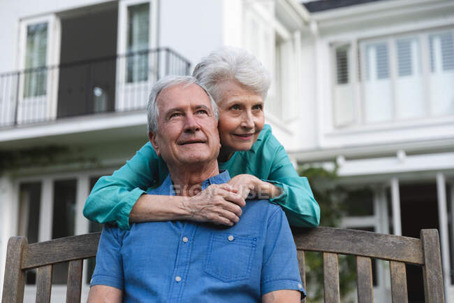 Feliz pareja de ancianos caucásicos jubilados en casa en el jardín fuera de su casa, el hombre sentado en un banco y la mujer de pie detrás abrazándolo, ambos mirando hacia otro lado y sonriendo, en casa juntos aislando durante coronavirus covidemic 19 - foto de stock