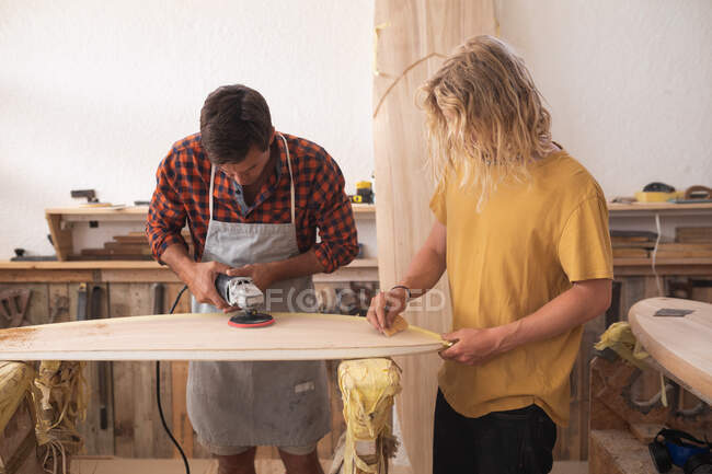 Dos surfistas caucásicos trabajando en su estudio y haciendo una tabla de surf de madera juntos, puliéndola y moldeándola con una lijadora. - foto de stock