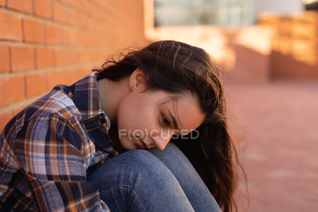 Бічний вид на Кавказьку сумну дівчину - підлітка, яка сидить на землі сама на шкільному подвір 