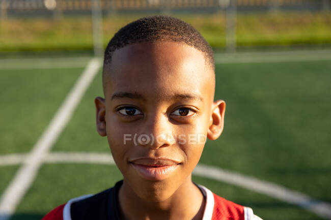 Portrait close up of a confident African American boy soccer player wearing a team strip, standing on a playing field in the sun, looking to camera and smiling — Stock Photo