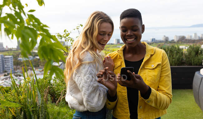 Front view of a Caucasian and an African American women hanging out on a roof terrace on a sunny day, using a smartphone and smiling — Stock Photo