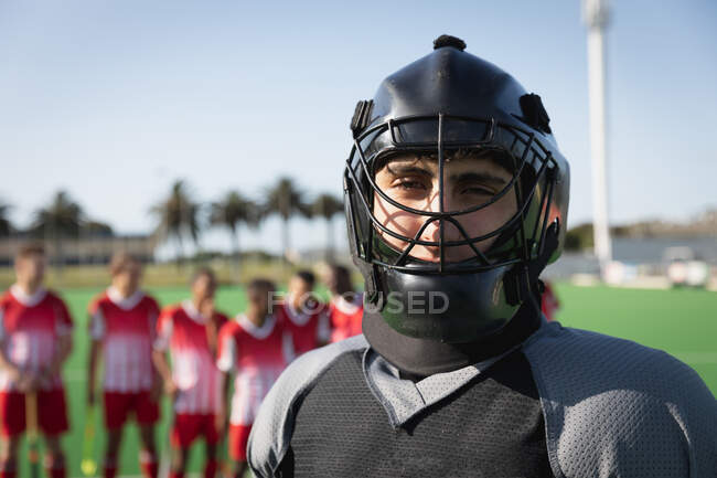 Portrait close up of a Caucasian male field hockey goalkeeper, wearing black hockey uniform and a helmet, standing on the pitch and looking to camera, with his teammates in the background on a sunny day — Stock Photo