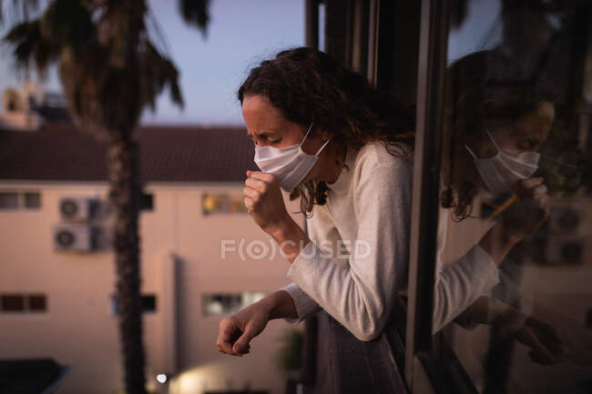 Mujer caucásica pasar tiempo en casa auto aislamiento y distanciamiento social en cuarentena de bloqueo durante coronavirus covid 19 epidemia, con una máscara facial contra covid19 coronavirus, mirando a través de la ventana y tos. - foto de stock