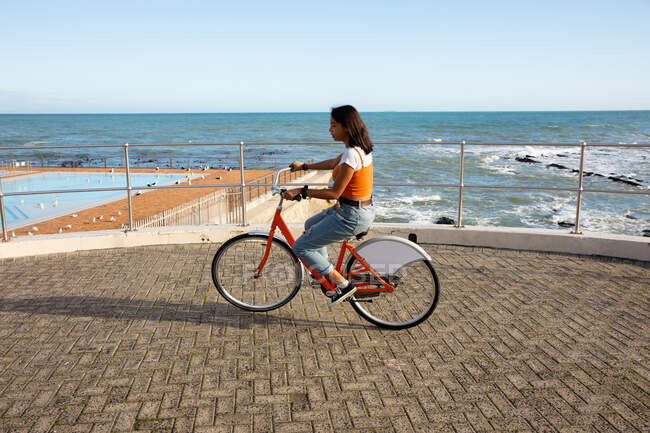 Seitenansicht eines gemischten Rennmädchens mit orangefarbenem Oberteil und Jeans, das an einem sonnigen Tag die Zeit genießt, mit dem Fahrrad auf der Strandpromenade zu fahren. — Stockfoto