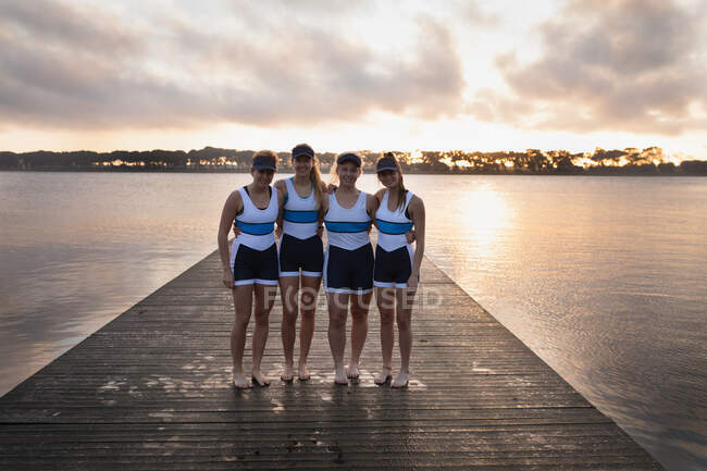 Portrait of a rowing team of four Caucasian women standing on a jetty with arms around each other, smiling to camera at sunrise — Stock Photo