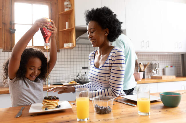 Front view of a young African American girl and her mother at home in the kitchen in the morning, sitting at the kitchen island, the girl pouring sauce on her pancakes and the mother laughing, with the father standing in the background cooking — Stock Photo