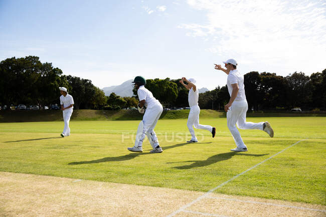 Seitenansicht einer multiethnischen Cricket-Mannschaft, die Weiße trägt und während eines Cricketspiels an einem sonnigen Tag durch das Spielfeld rennt. — Stockfoto