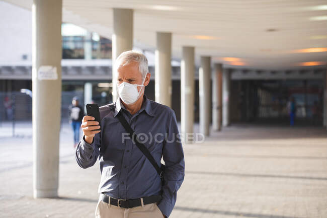 Hombre caucásico mayor por las calles de la ciudad durante el día, con una máscara facial contra el coronavirus, covid 19, usando un teléfono inteligente. - foto de stock