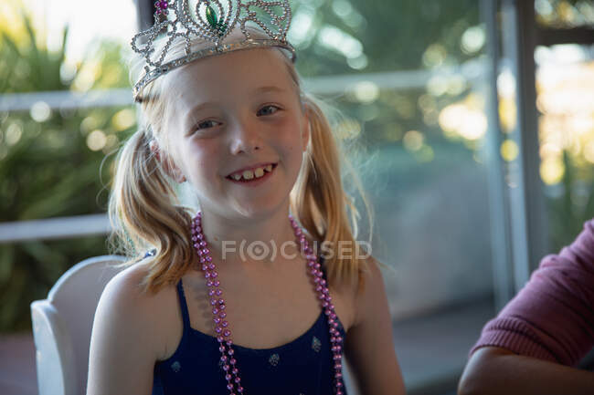 Vista frontal de uma menina caucasiana desfrutando de tempo livre com sua mãe em casa, sentada em uma mesa na sala de estar, vestindo coroa e colar, sorrindo para a câmera — Fotografia de Stock