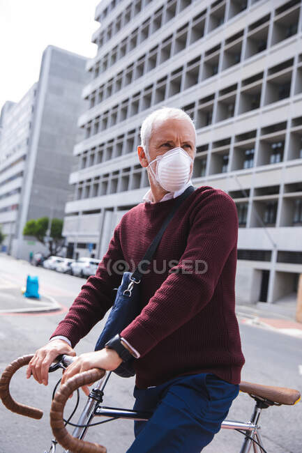 Uomo anziano caucasico in giro per le strade della città durante il giorno, indossando una maschera contro il coronavirus, covid 19, in sella alla sua bicicletta. — Foto stock