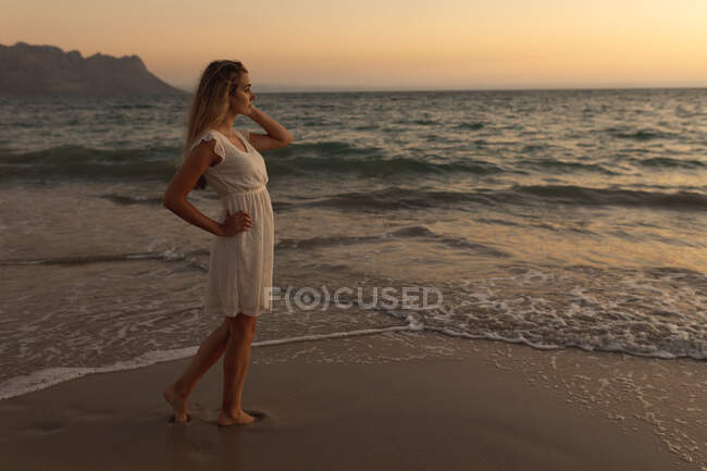 Mujer caucásica vistiendo un vestido blanco de pie descalzo en una playa al atardecer, mirando al mar, relajándose durante unas vacaciones activas en la playa - foto de stock