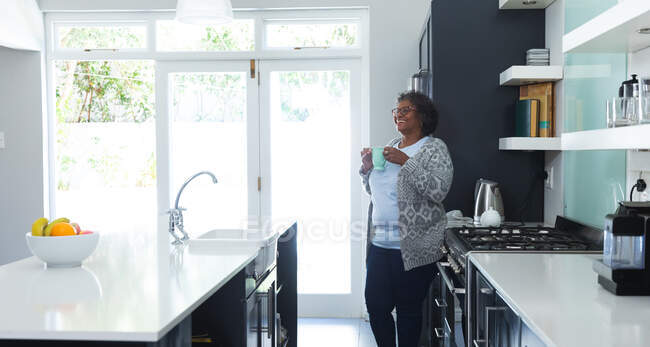 Mulher de raça mista sênior desfrutando de seu tempo em casa, distanciamento social e auto-isolamento em quarentena, de pé em sua cozinha, segurando uma xícara e sorrindo — Fotografia de Stock