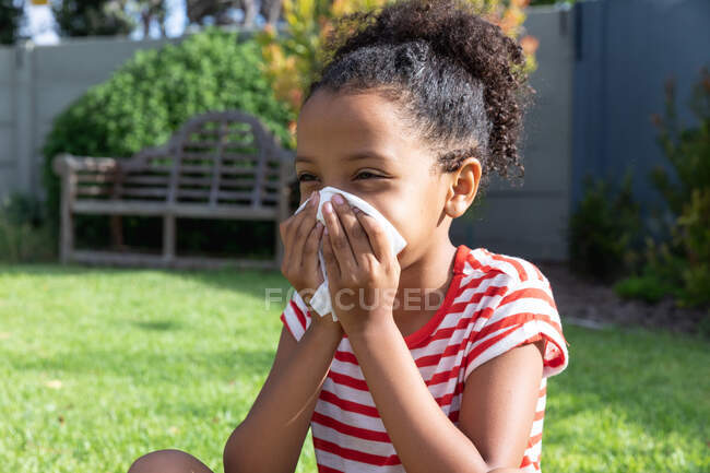African American girl social distancing at home during quarantine lockdown, wearing a stripped t-shirt, covering her nose with a tissue while sneezing, in a garden on a sunny day. — Stock Photo