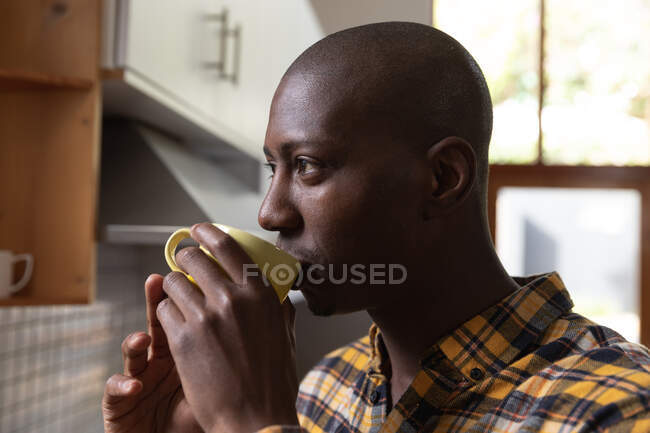Vista laterale da vicino di un uomo afroamericano a casa, in piedi in cucina a bere una tazza di caffè e distogliere lo sguardo — Foto stock
