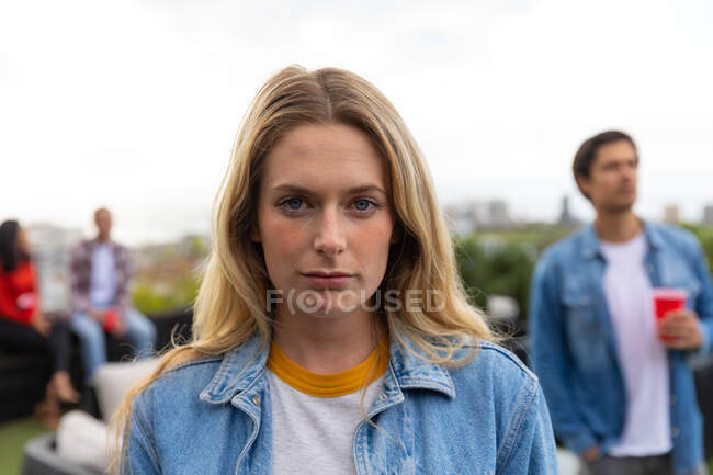 Portrait of a Caucasian woman hanging out on a roof terrace on a sunny day, looking at camera, with people talking in the background — Stock Photo
