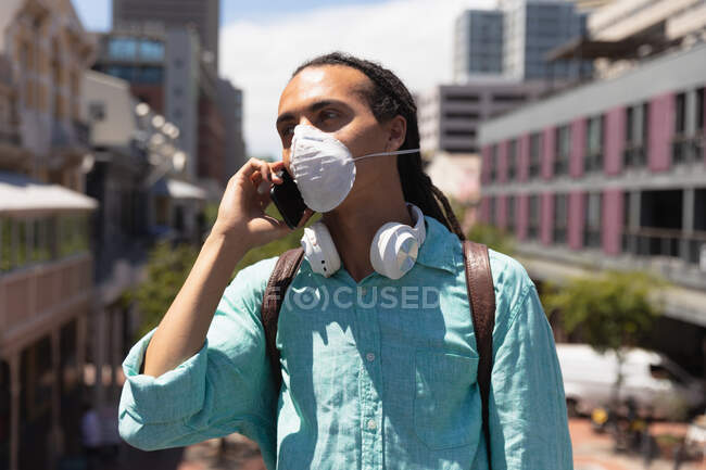Vista frontal de un hombre de raza mixta con rastas largos en la ciudad en un día soleado, parado en la calle con una máscara de coronavirus y usando un teléfono inteligente - foto de stock