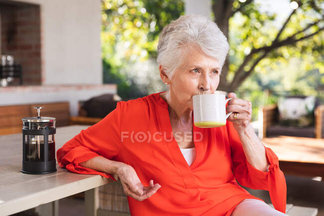 Eine glückliche Rentnerin aus dem Kaukasus, die an einem sonnigen Tag zu Hause im Garten vor ihrem Haus sitzt, an einem Tisch mit einer Tasse Kaffee sitzt, wegschaut und lächelt, sich während der Coronavirus-Pandemie selbst isoliert 19 — Stockfoto