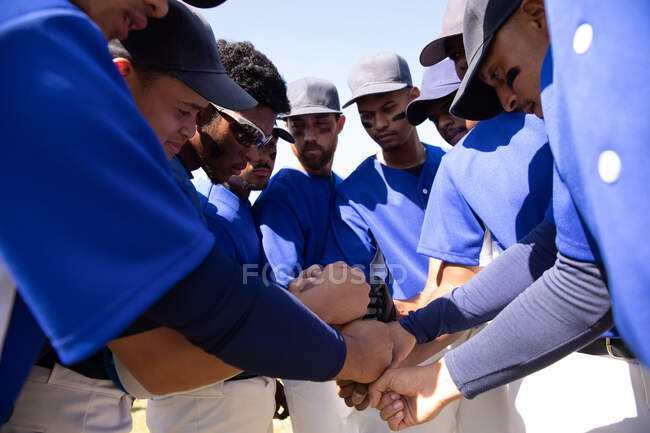 Vue latérale d'un groupe multi-ethnique de joueurs de baseball masculins, se préparant avant un match, se blottissant par équipe, empilant les mains, se motivant mutuellement — Photo de stock