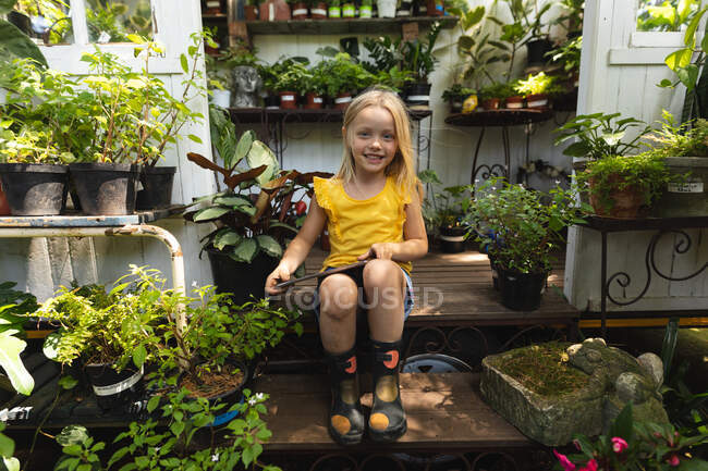 Portrait of a Caucasian girl with long blonde hair enjoying time in a sunny garden, sitting in the doorway of a greenhouse, using a tablet computer, looking at camera and smiling — Stock Photo
