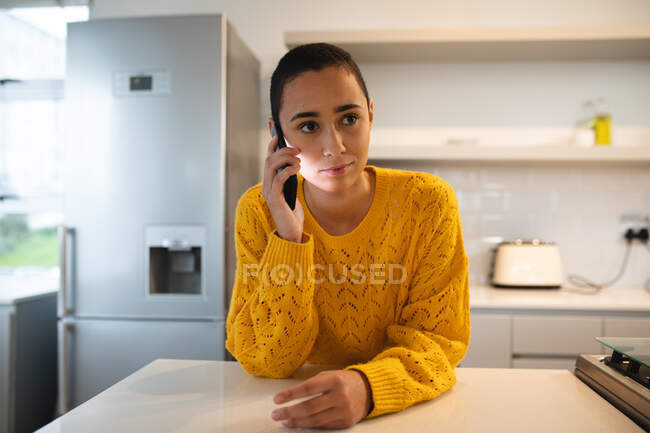 Вид спереду змішаної раси жінка з коротким волоссям, що розслабляється вдома, стоїть на кухні, спираючись на стільницю, має телефонний дзвінок на смартфон — стокове фото